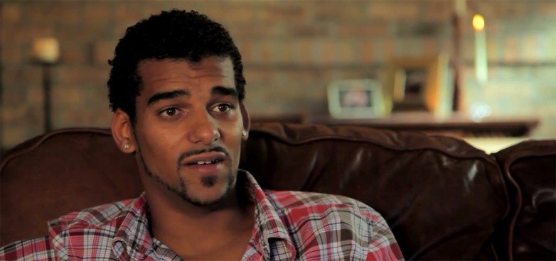Young man with curly hair and a goatee in a plaid shirt speaking on a leather couch