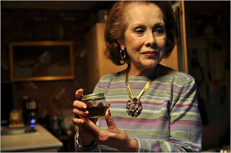 Elderly woman in a striped sweater holding a small jar of green cannabis oil in a kitchen