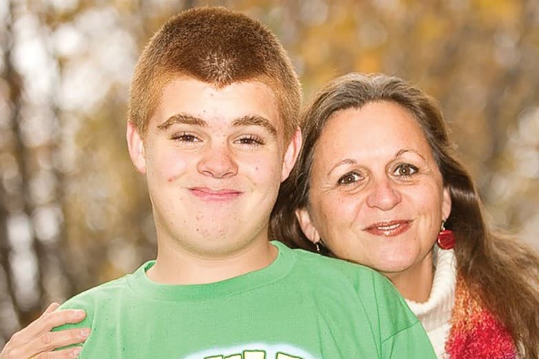 Smiling teenage boy in a green shirt beside a woman outdoors among autumn trees