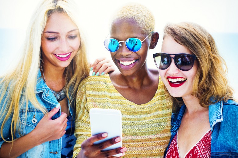 Three young women in sunglasses smiling at a smartphone outdoors in bright sunlight