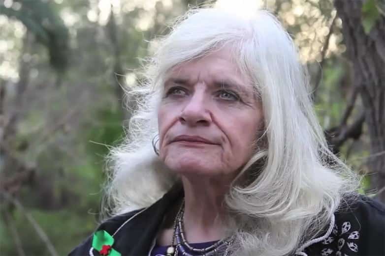 Older woman with long white hair and beaded necklace outdoors among trees