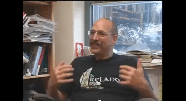 Man in an 'Ireland' t-shirt gesturing while talking in a cluttered office