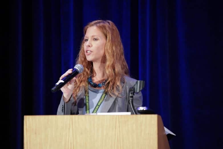 Red-haired woman speaking into a microphone at a lectern before a blue curtain