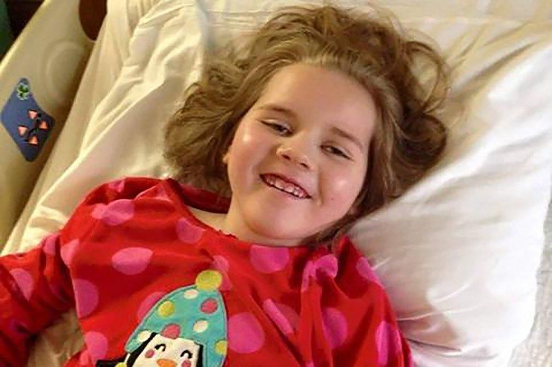 Smiling young girl with tousled hair lying against a pillow in a hospital bed, red pajamas