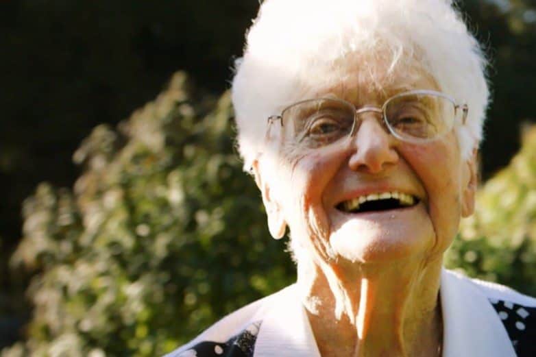 Elderly woman with white hair and glasses laughing outdoors with greenery behind her