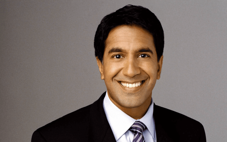 Sanjay Gupta headshot smiling in a dark suit and purple striped tie against a gray backdrop