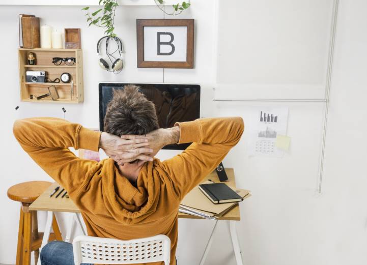 Man in an orange hoodie leaning back with hands behind his head at a tidy home-office desk
