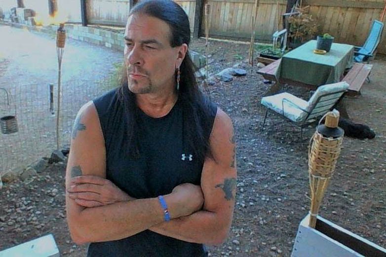 Man with long dark hair and crossed arms standing in a backyard with tiki torches