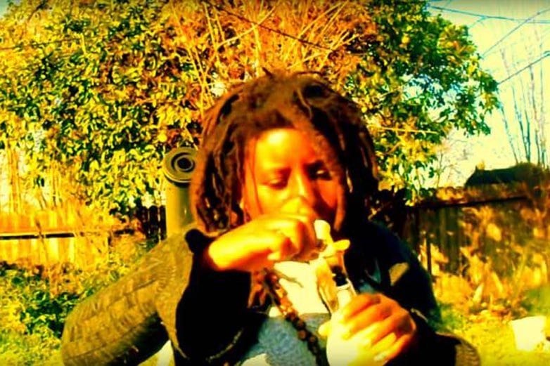 Person outdoors exhaling smoke while holding a glass water pipe, sepia-toned backyard