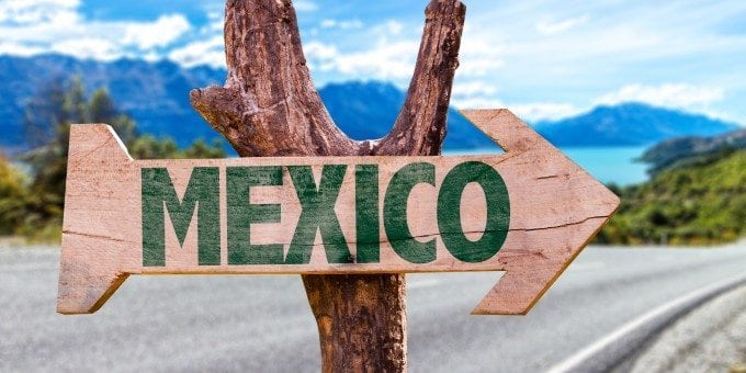 Wooden arrow sign reading MEXICO pointing right, with a highway, lake and mountains behind